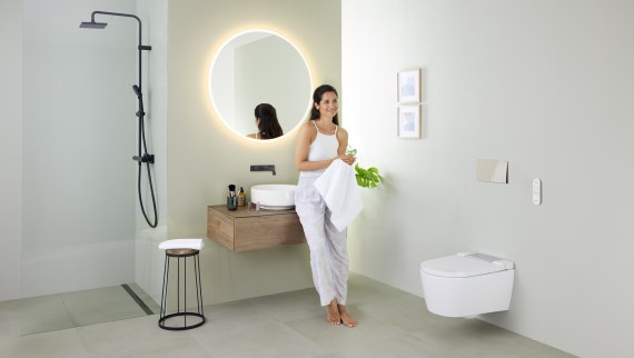 Geberit AquaClean Sela otomatik taharet sistemi ve Geberit VariForm lavabo ve mobilyaların bulunduğu banyoda lavaboya yaslanan kadın
